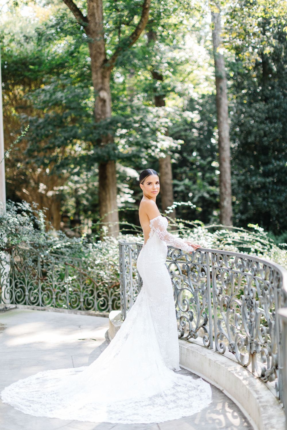 Elegant bride in lace mermaid gown poses on stone bridge surrounded by lush garden trees and ornate iron railings.