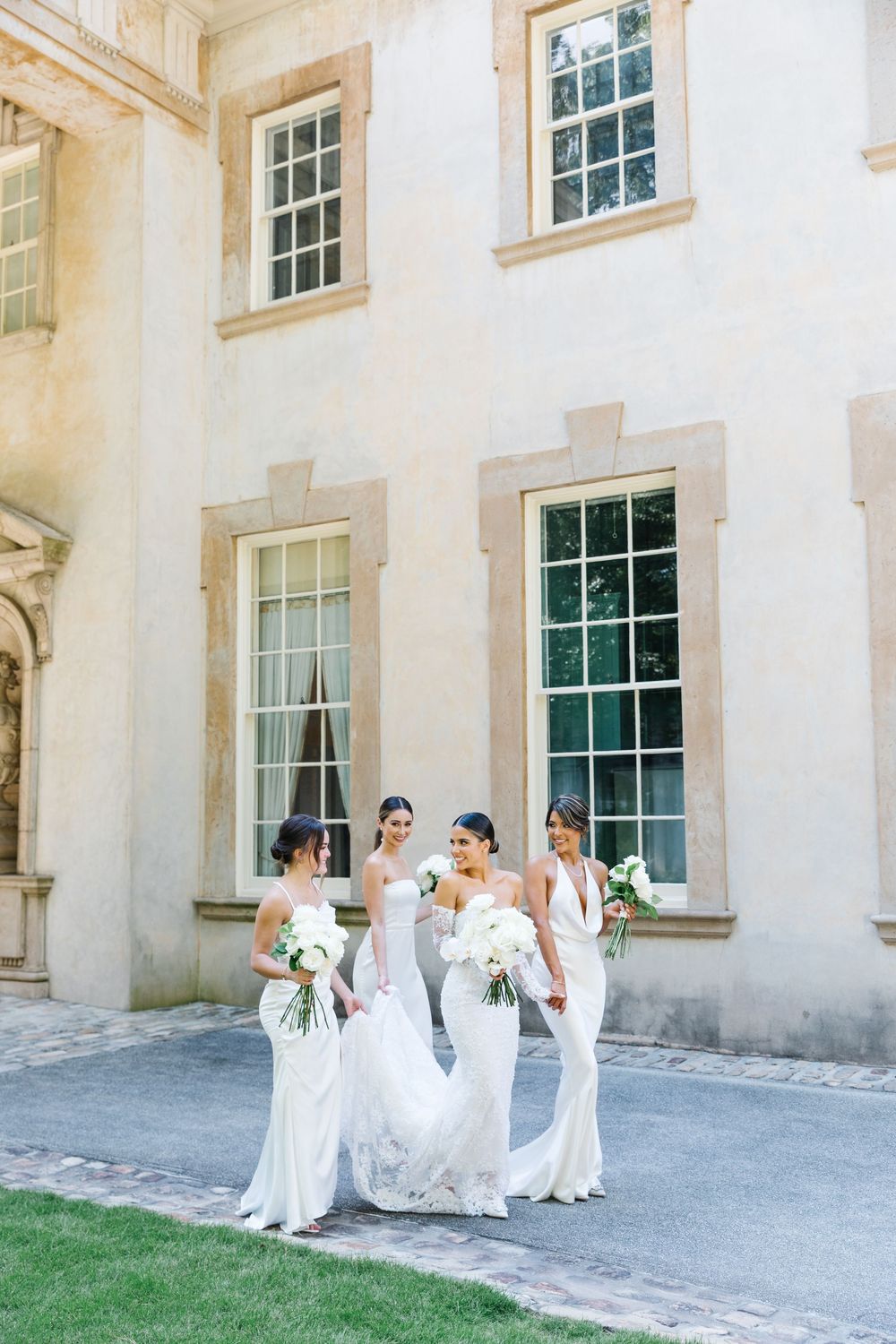 A group in elegant white dresses stands together on a sunny day outside a historic building with tall windows.