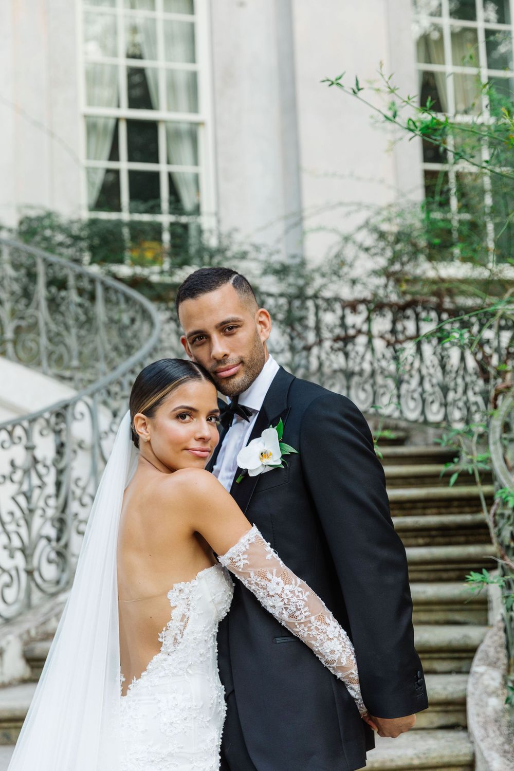A newlywed couple poses elegantly on grand stone steps with ornate iron railings and white columns in the background.