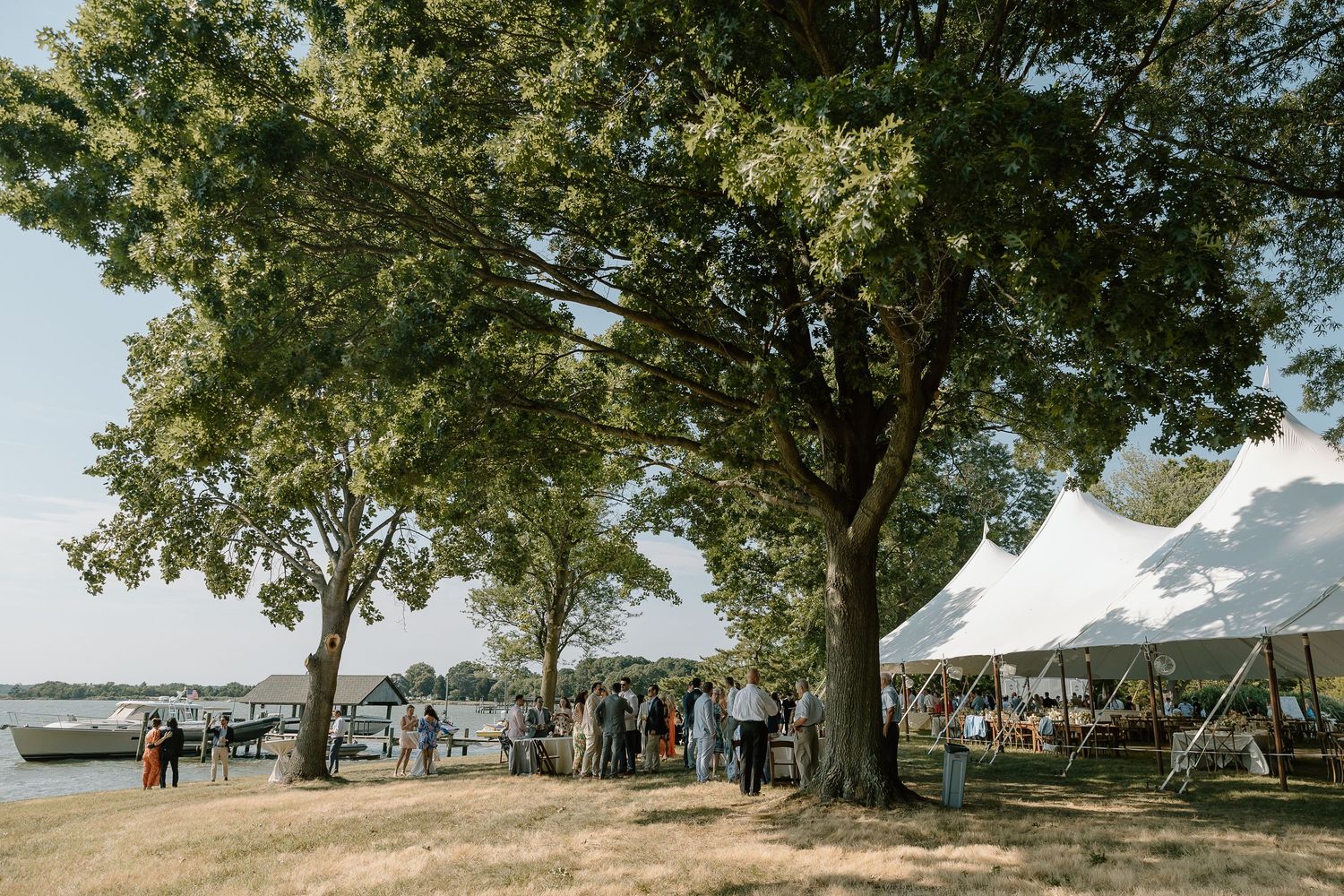 Outdoor wedding reception under tent with large trees and waterfront views.