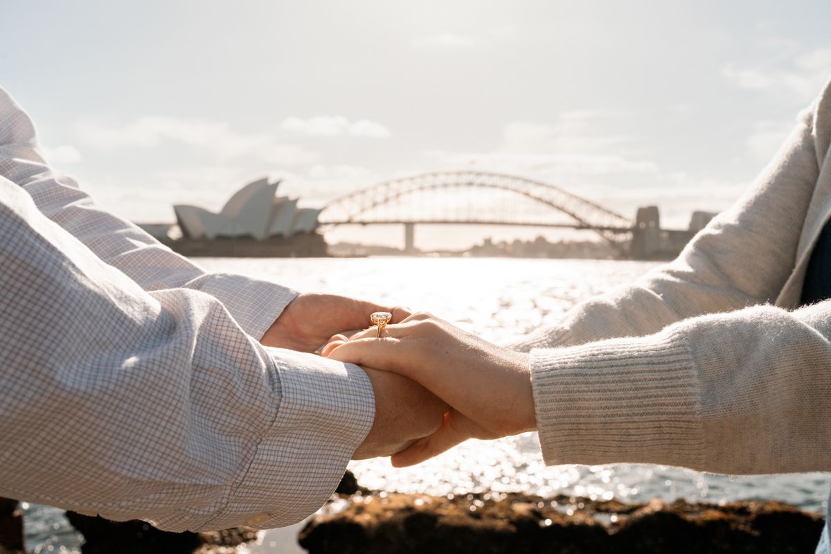 close up photo of man and woman holding hands looking at each other after a proposal background is Sydney opera house and the harbour bridge