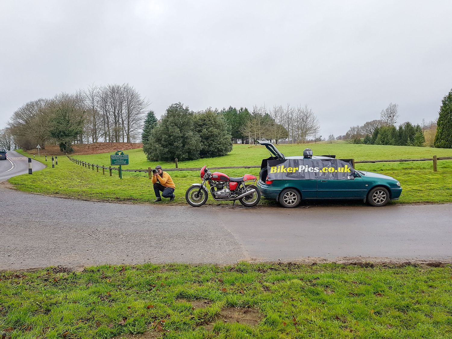 A motorcycle and car are parked on a road alongside a green grassy field with trees in the background.
