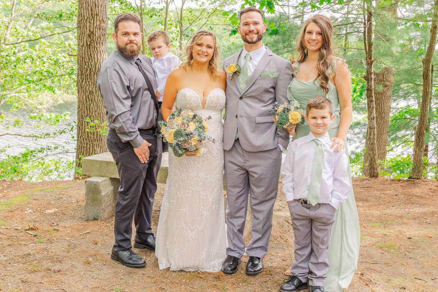Wedding party stands together on a wooded path with the couple in white and gray attire surrounded by trees.