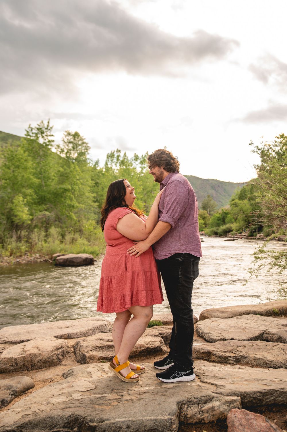 A couple shares an intimate moment while standing on rocky terrain near a rushing river with mountains in the background.