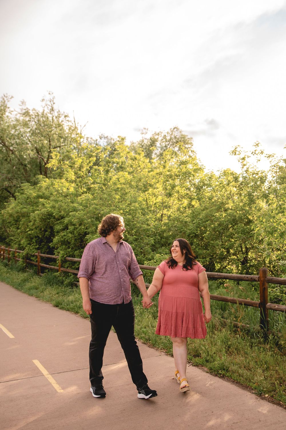 A couple holds hands while walking along a paved trail bordered by a wooden fence and lush greenery during sunset.