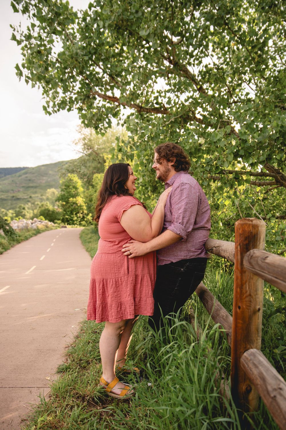 A romantic couple embraces near a wooden fence on a paved trail surrounded by green foliage and hills.