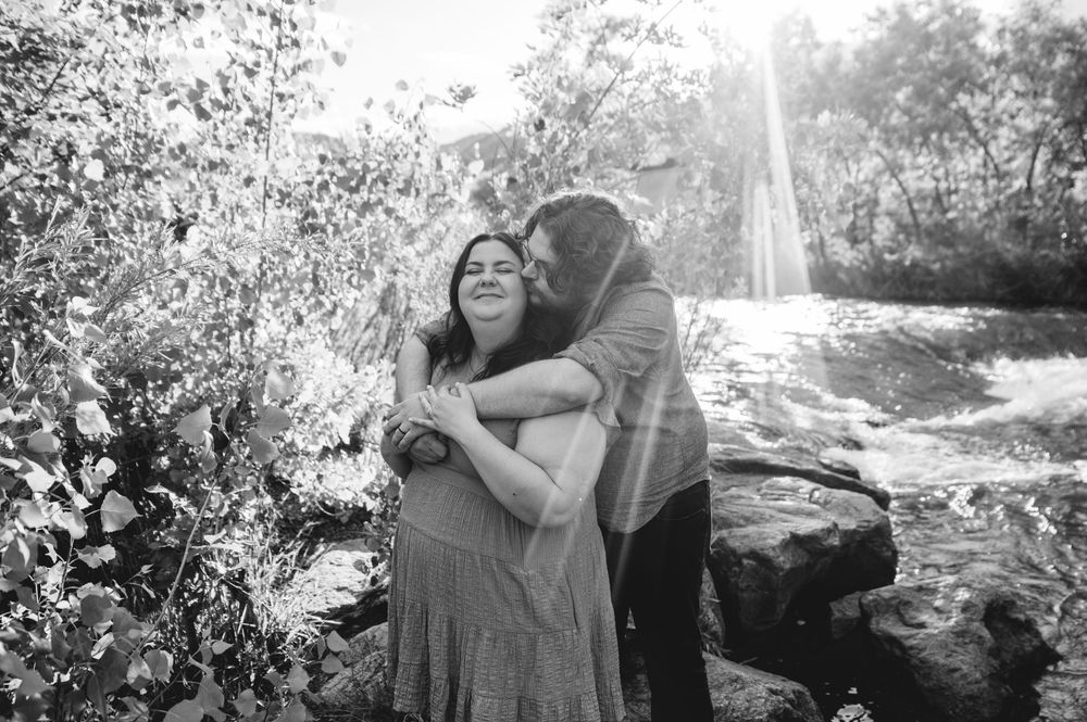 A couple embraces in black and white by a sunlit waterfall surrounded by rocks and foliage.