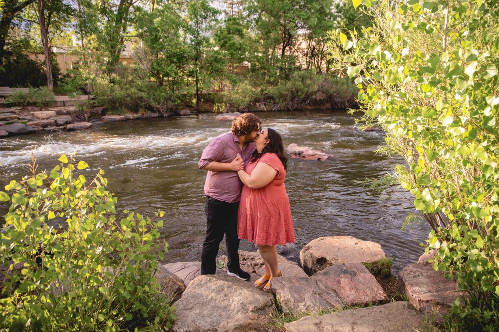 A romantic couple embrace while standing on large rocks beside a flowing river surrounded by spring foliage.