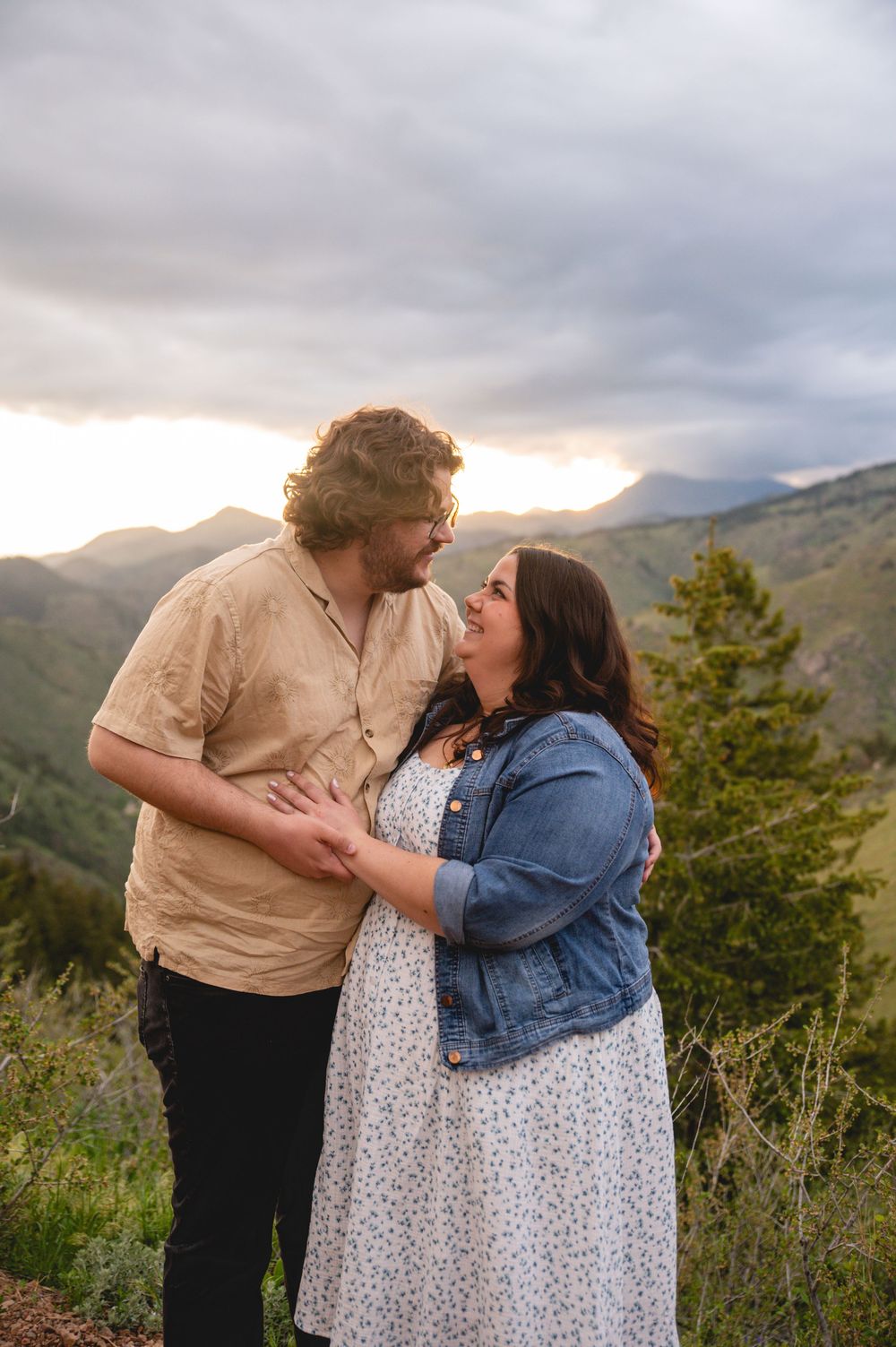 A couple shares an intimate moment on a scenic mountain overlook at sunset with dramatic cloudy skies in the background.