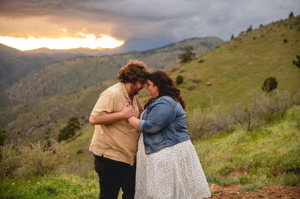Two people share an intimate moment on a grassy hillside at sunset with mountains in the background.