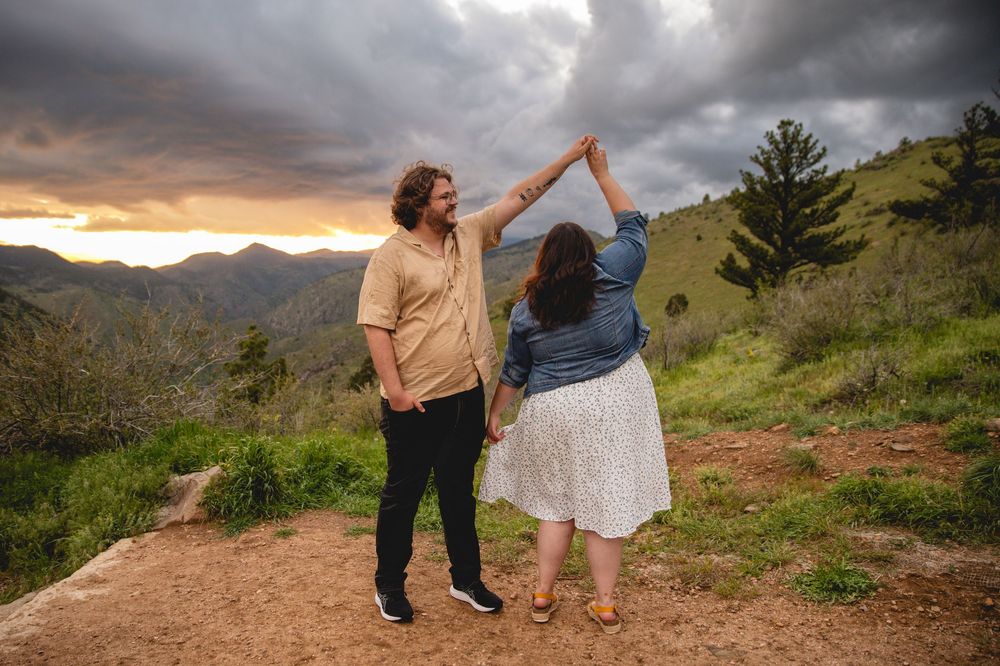 Two people dance together on a mountain trail at sunset with dramatic cloudy skies and scenic mountain views behind them.