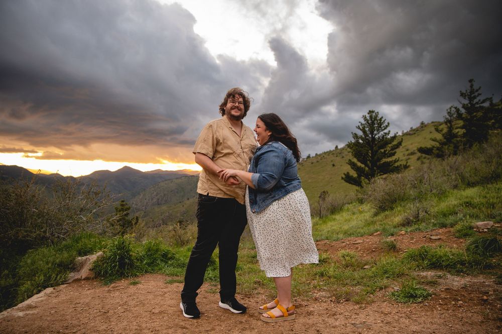 Two people stand together on a mountain overlook at sunset with dramatic storm clouds overhead in the background.