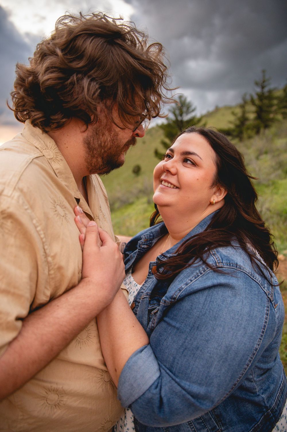 A couple shares a tender romantic moment outdoors with mountains and evergreen trees in the background.