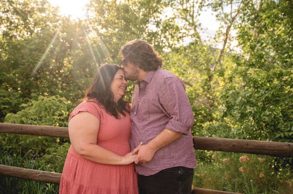 A couple shares a romantic moment together on a rustic wooden bridge surrounded by lush green trees during sunset.
