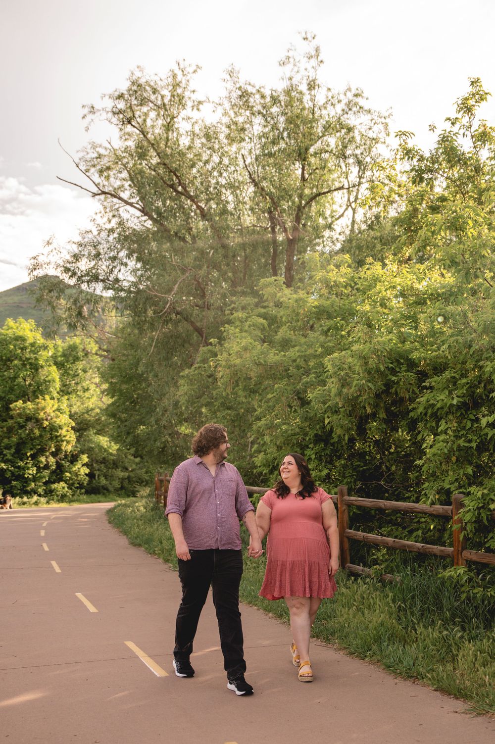A couple walks together on a paved nature trail lined with lush green trees and a wooden railing at sunset.