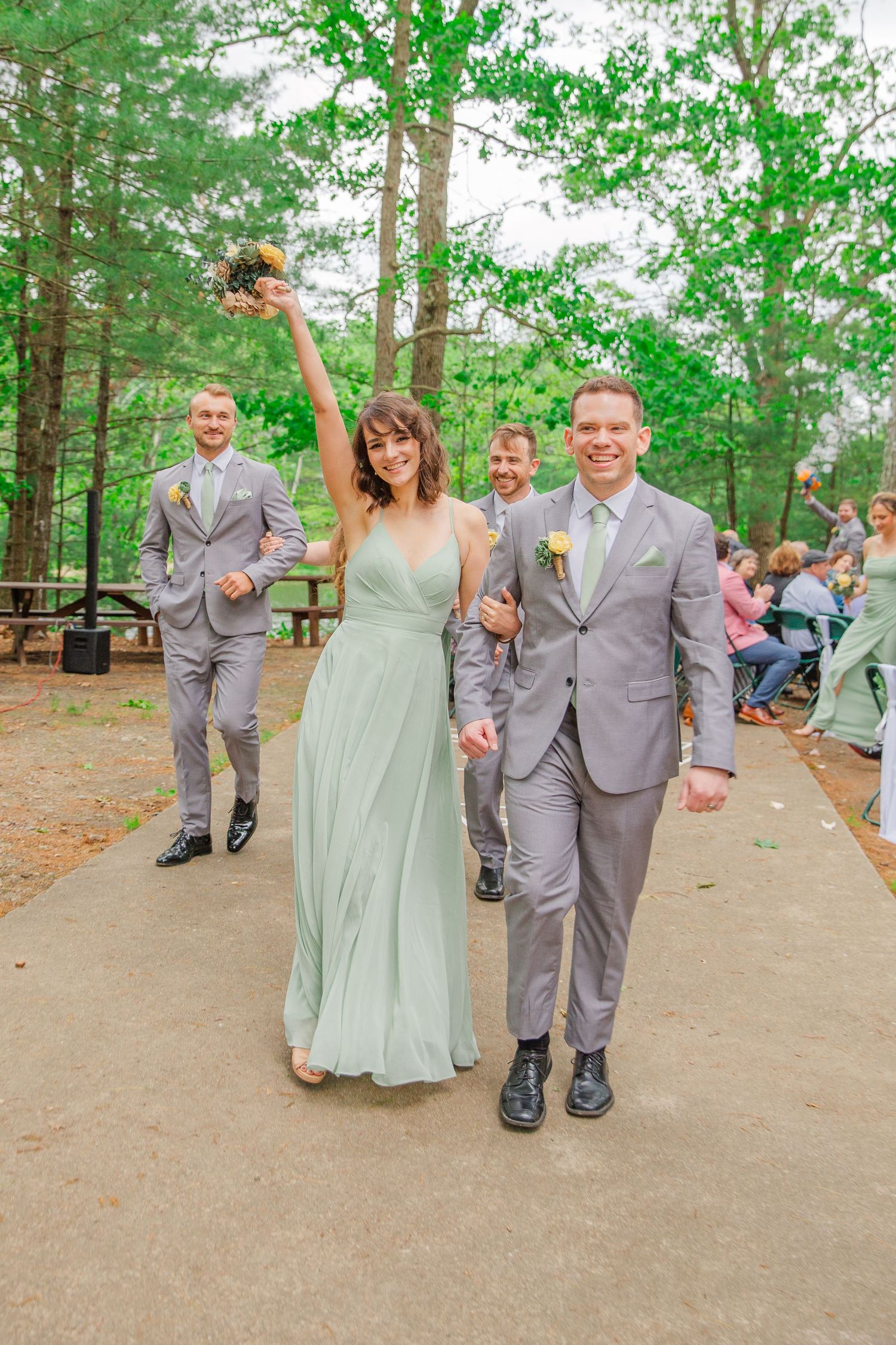 A joyful celebration in a wooded outdoor wedding venue with celebrants wearing sage gray suits and a mint green dress.