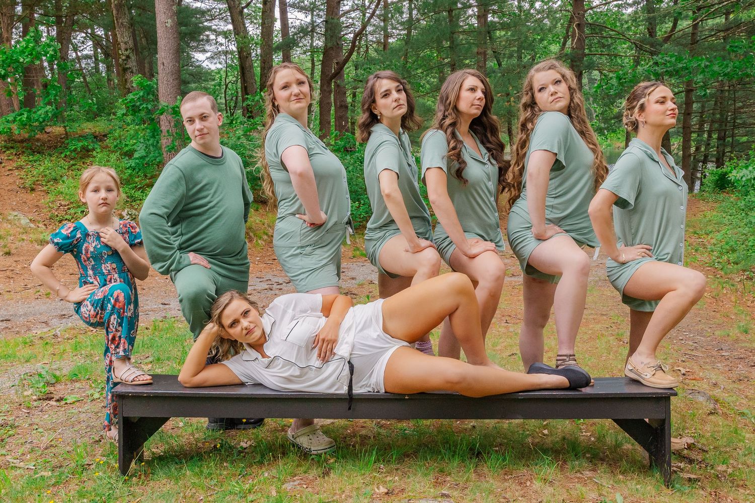 A group of people pose playfully together on wooden benches in a forested outdoor setting.