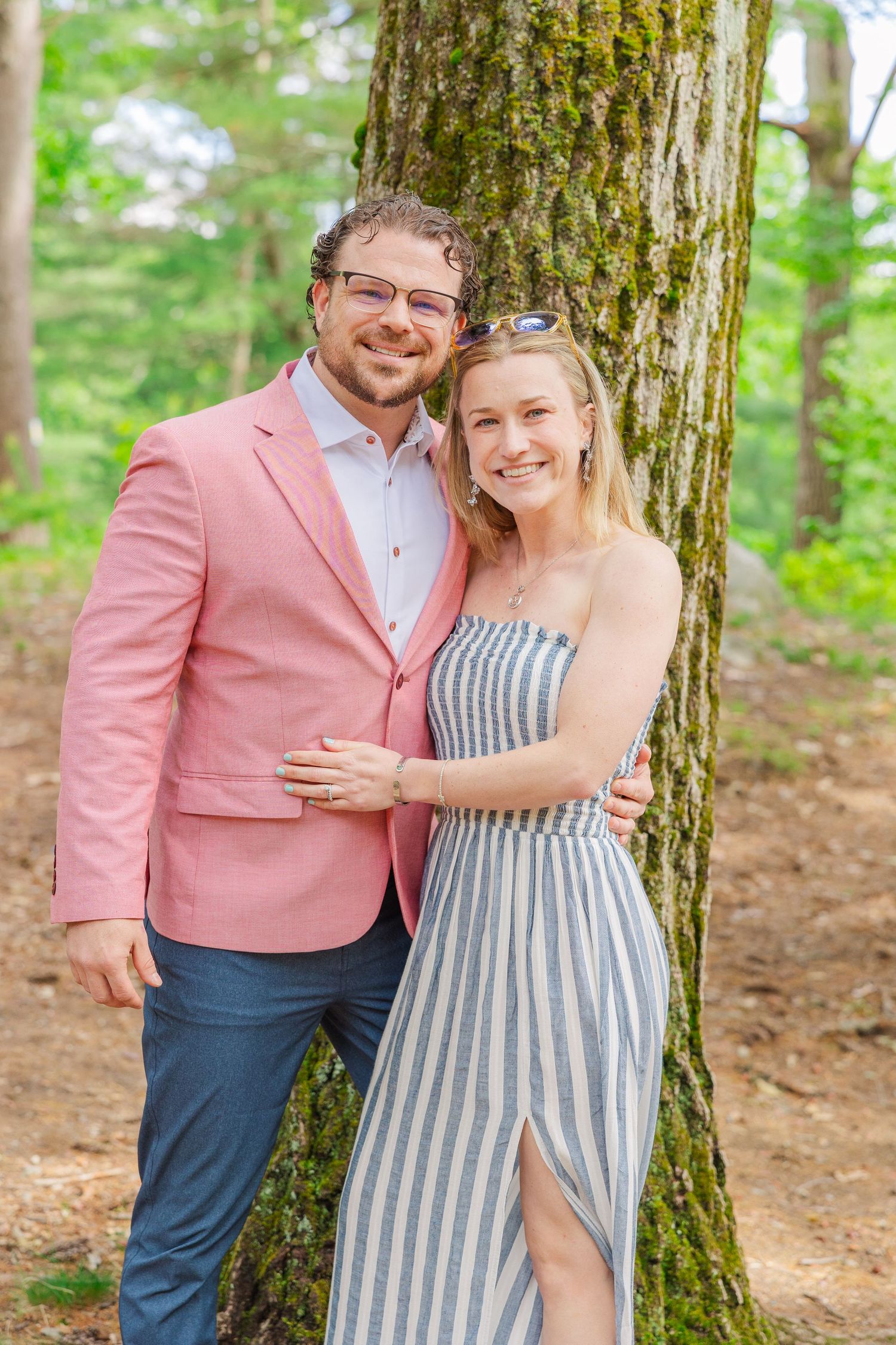 A couple poses together outdoors by a mossy tree trunk, wearing a pink blazer and striped sundress in a forest setting.