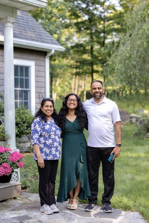 Three people stand together smiling on a stone patio outside a house with pink flowers and greenery in the background.