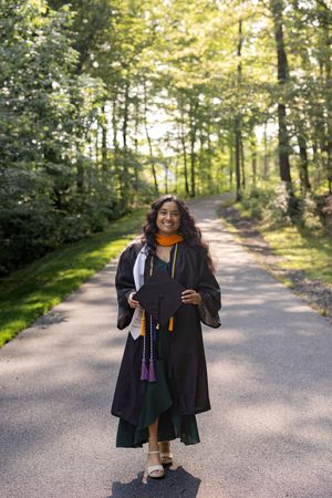Graduate in black cap and gown smiling on sunny tree-lined path during outdoor graduation photoshoot.