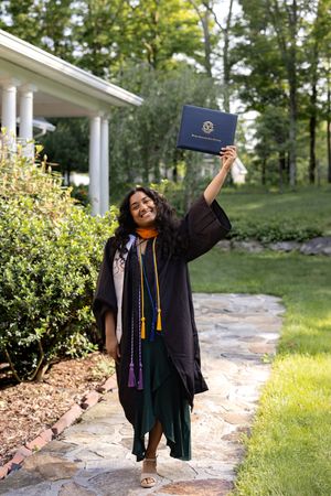 Graduate in black cap and gown holds diploma high while standing on stone path surrounded by greenery.
