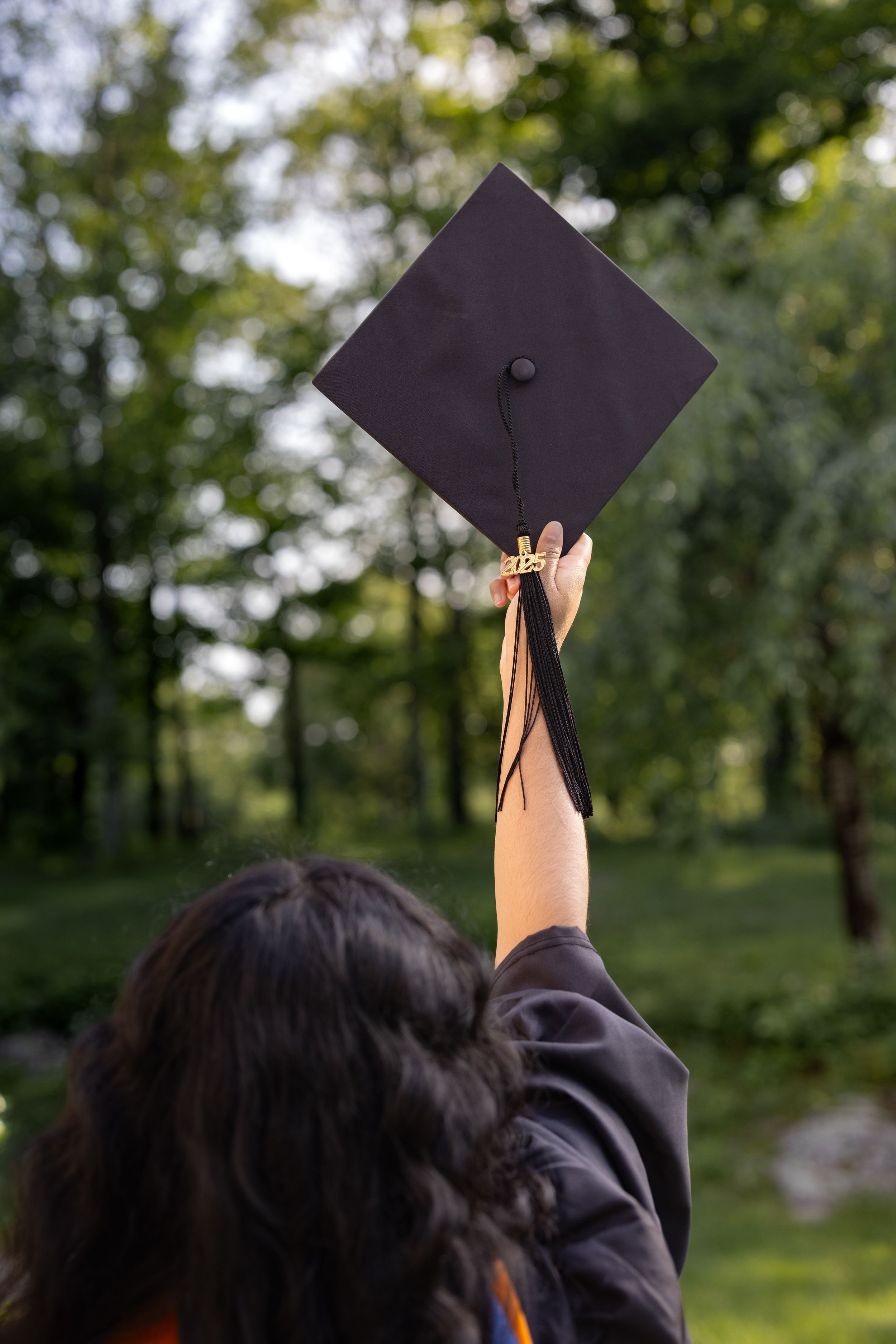 Graduate holds up black mortarboard graduation cap against blurred green forest backdrop in celebration.