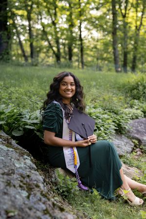 Graduate in dark green dress sits on rock in forest setting holding graduation cap with honors cords.
