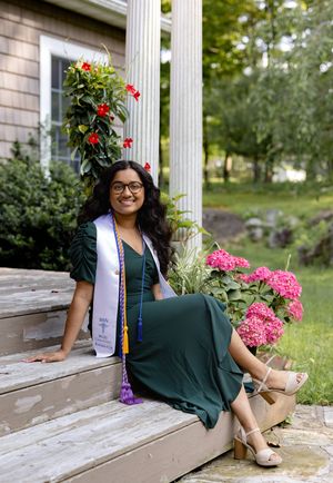 Graduate in emerald green dress and stole sits on wooden steps by columns with pink and red flowers blooming nearby.