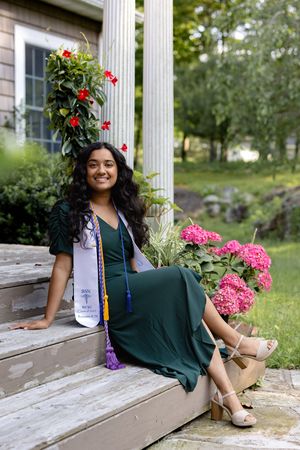 Graduate in green dress and floral crown sits on wooden bench in outdoor garden setting for graduation photos.