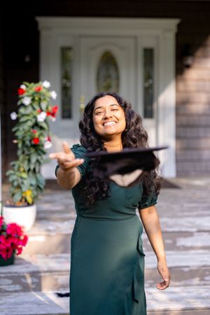 A joyful person in an emerald dress tosses their graduation cap in front of a decorated entrance.