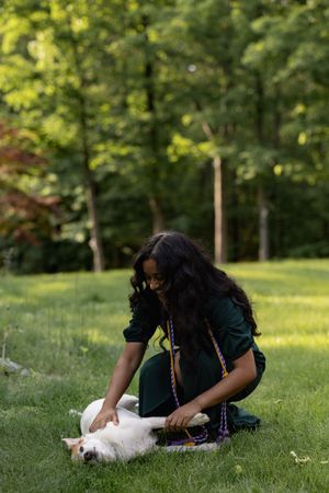 A person sitting in a grassy park pets a white cat laying on its back in warm sunlight surrounded by trees.