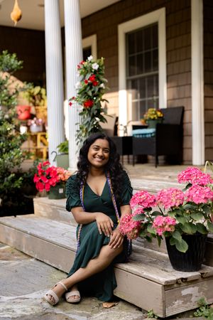 A person sits on wooden porch steps surrounded by vibrant pink flowers and climbing roses near white columns.