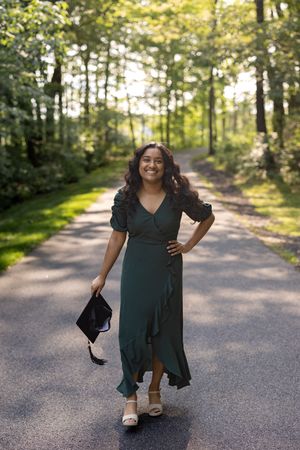 Someone in a flowing dark green wrap dress poses on a sunlit forest path with sandals and a handbag.