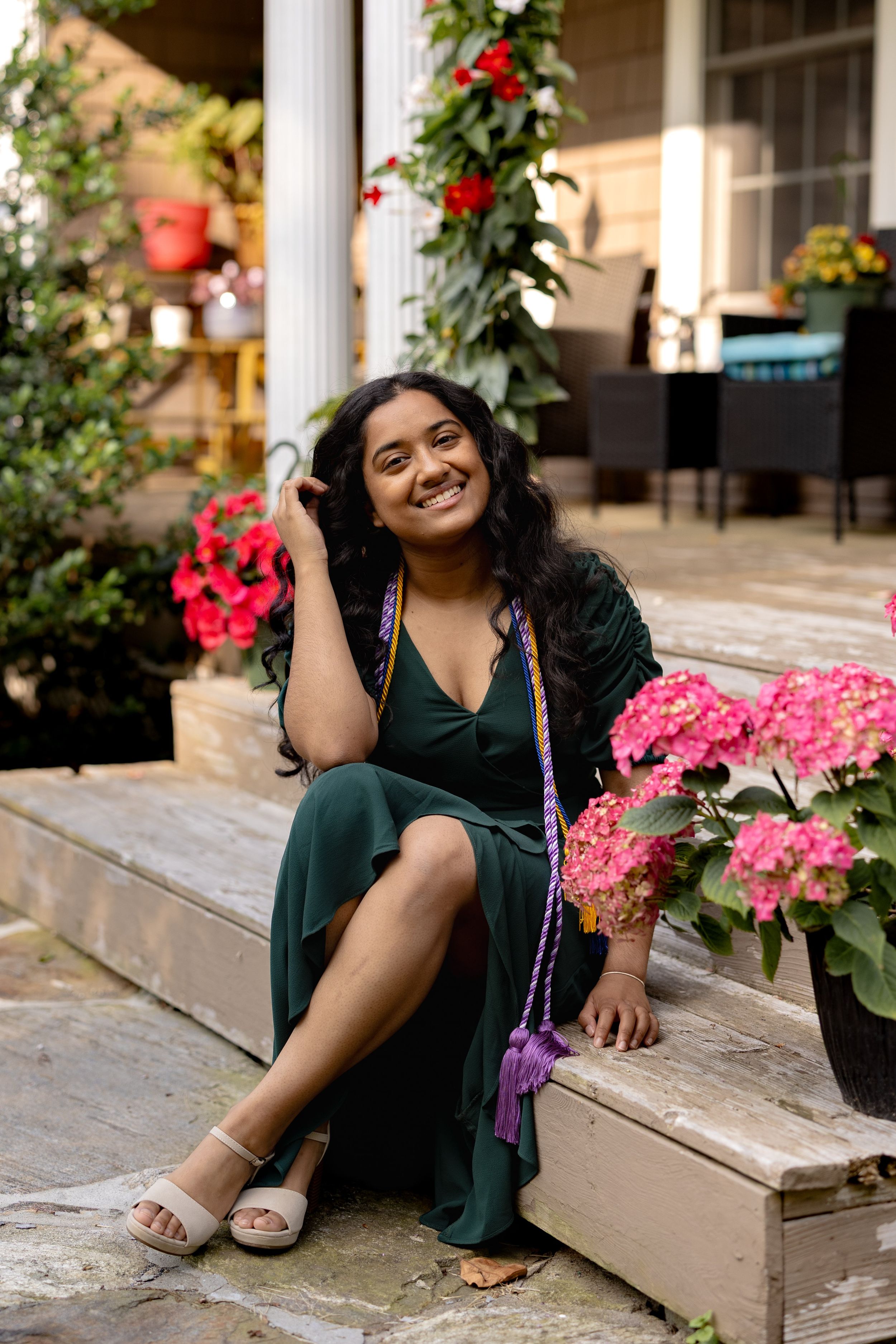 Woman in an elegant green dress sits on porch steps surrounded by vibrant pink hydrangeas and climbing red vines.