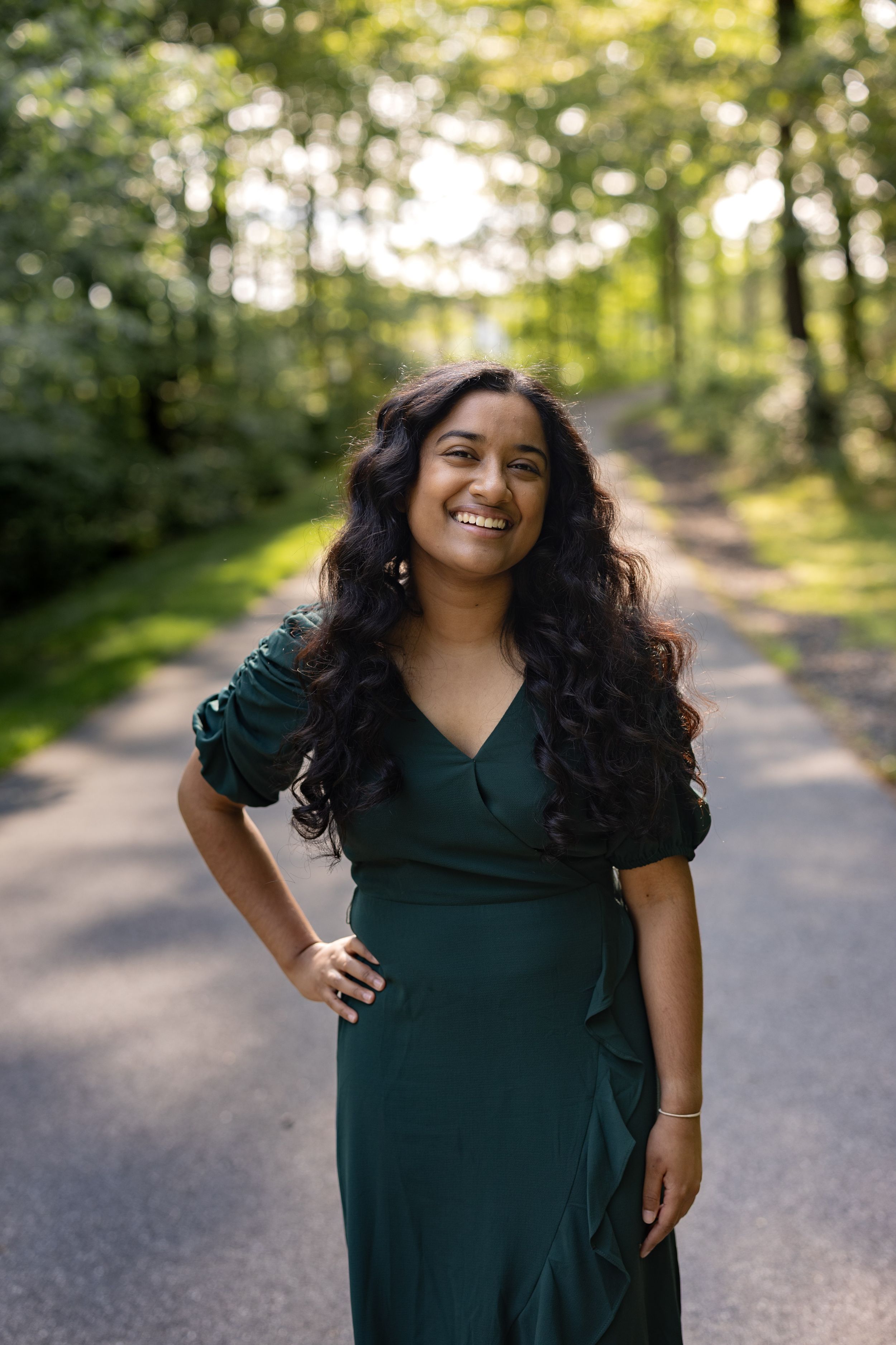 A cheerful individual in a dark green wrap dress poses on a sunlit forest path with flowing dark hair.