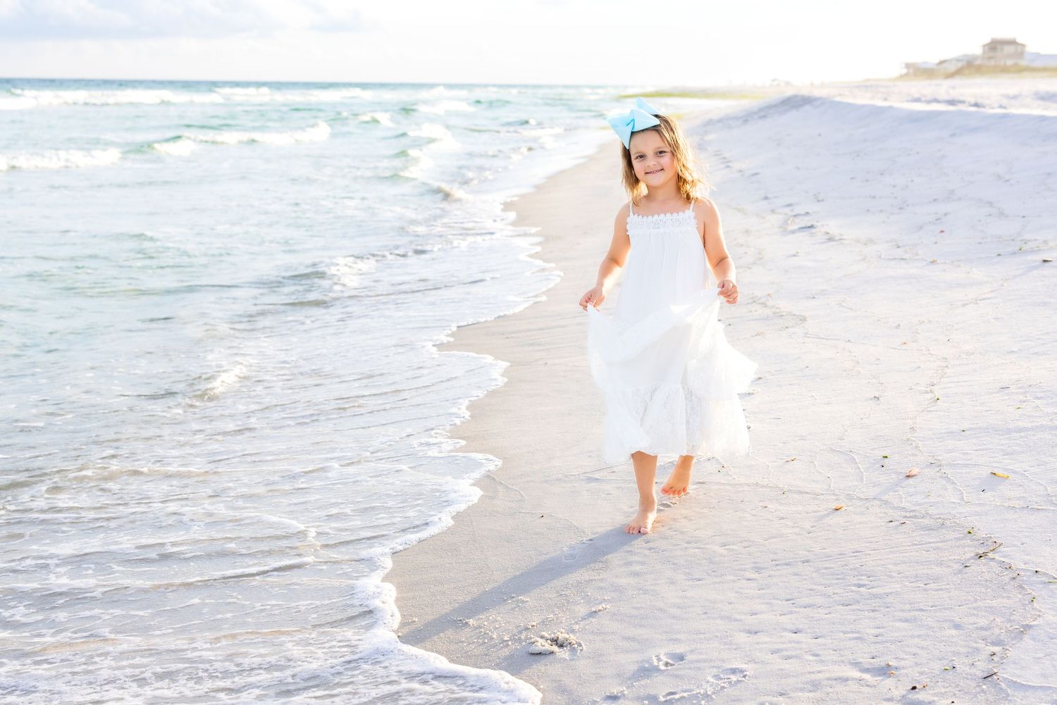 Woman in a white flowing dress walking along a serene beach shoreline at sunset while waves crash nearby.