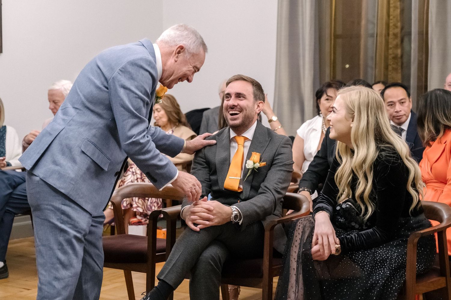 People in formal attire interact and laugh during an indoor social gathering at an elegant venue.