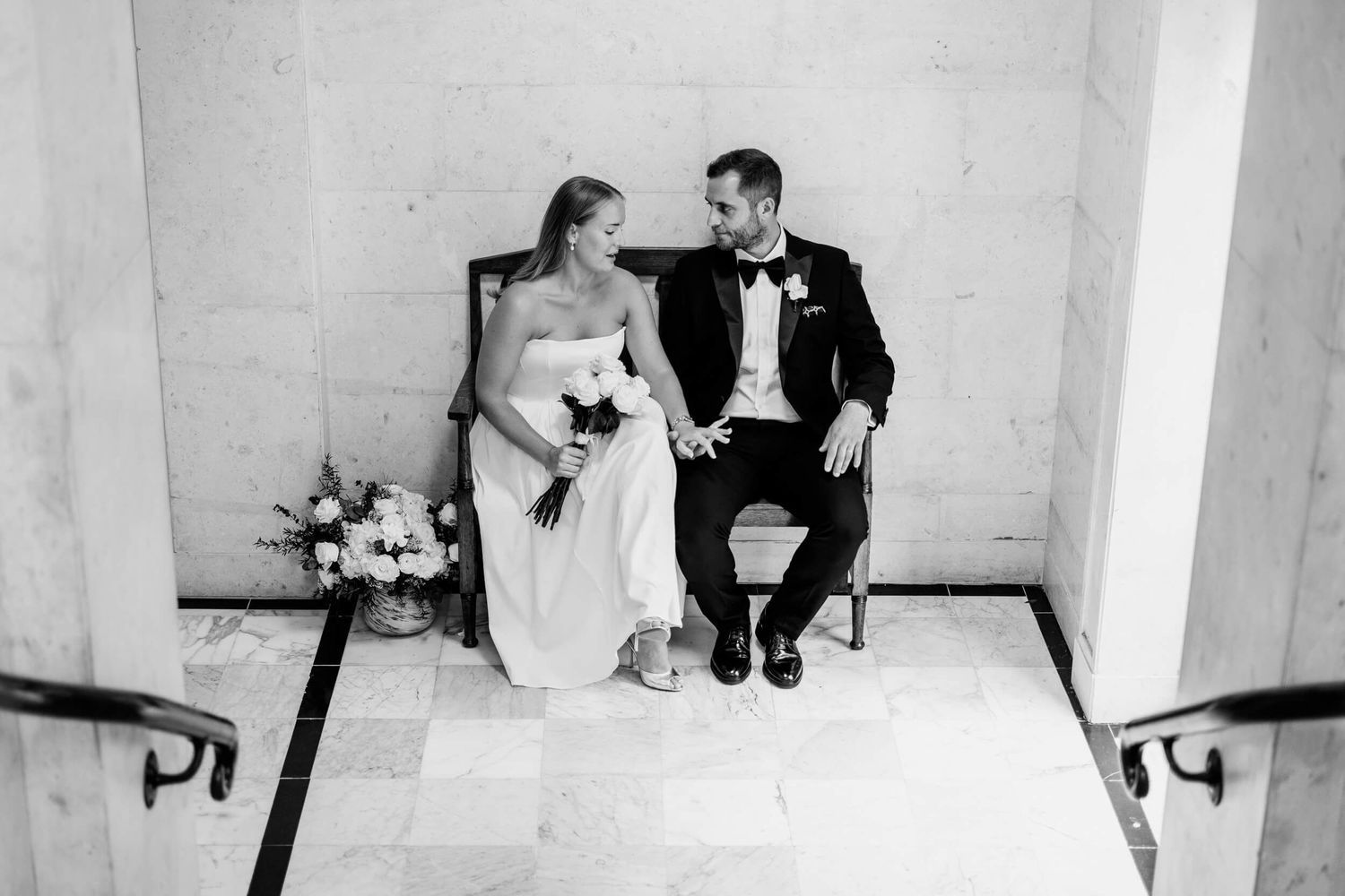 A wedding couple sits together on white marble steps in an elegant black and white photograph.