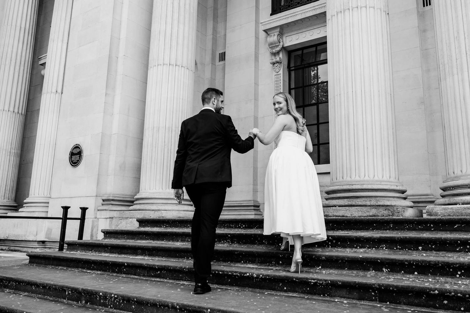 Black and white photo of a couple ascending grand stone steps between classical columns at a historic building.