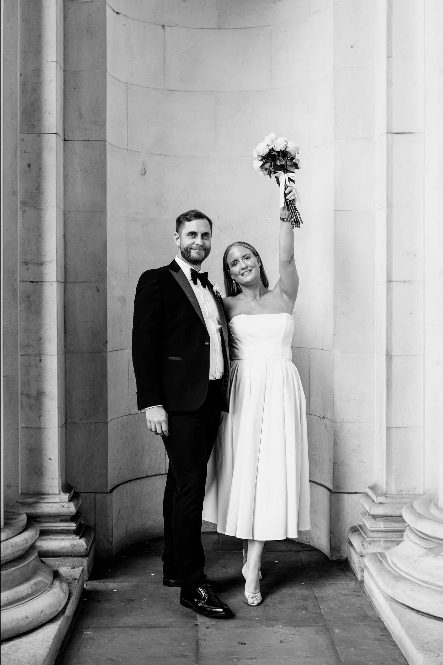 A black and white wedding photo showing a couple in formal attire posing elegantly between classic stone columns.