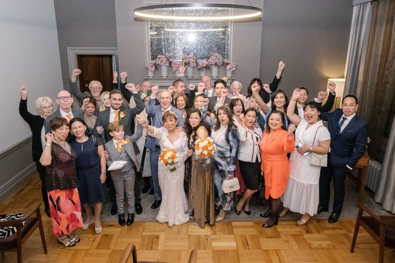 Large group photo of wedding guests and couple posing together in a reception venue with chandelier.