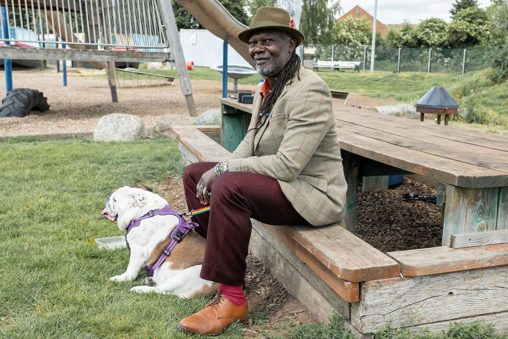 Man in hat and blazer sitting on wooden bench outdoors with a dog resting nearby.