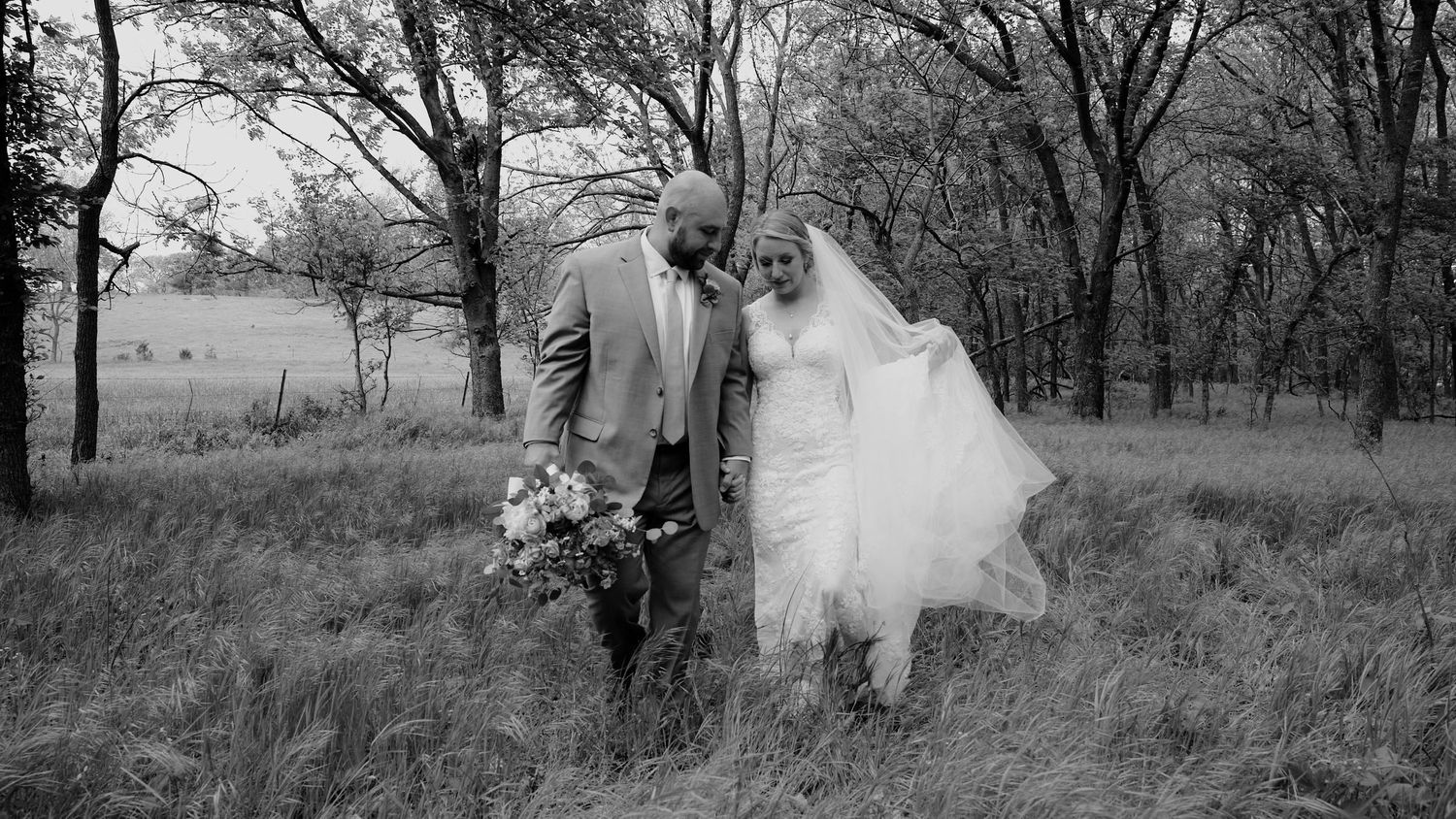 A couple walks hand in hand through a lush green meadow with trees while holding a vibrant floral bouquet.