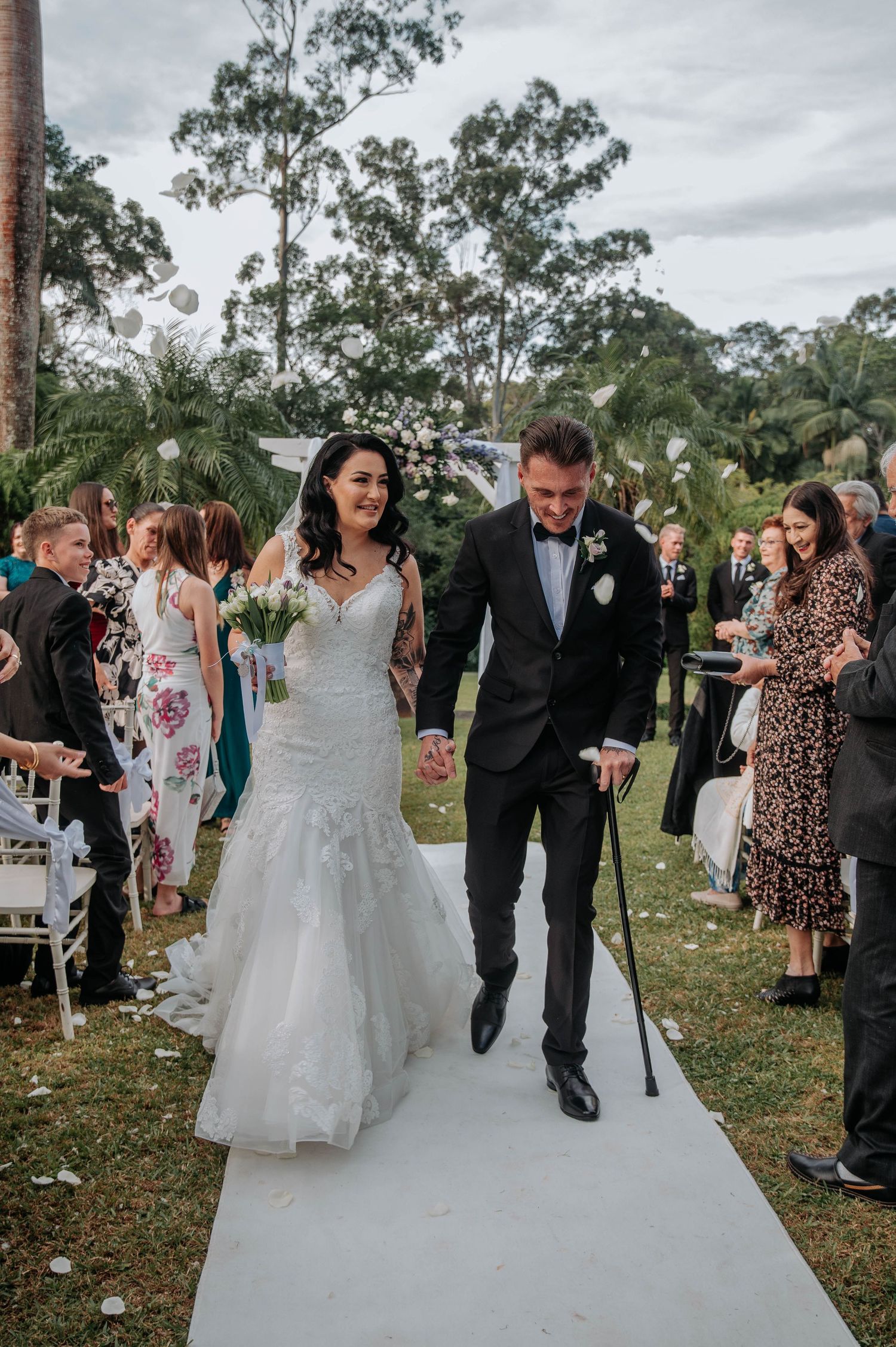 Wedding guests line a pathway tossing flower petals as newlyweds walk together after their outdoor ceremony.
