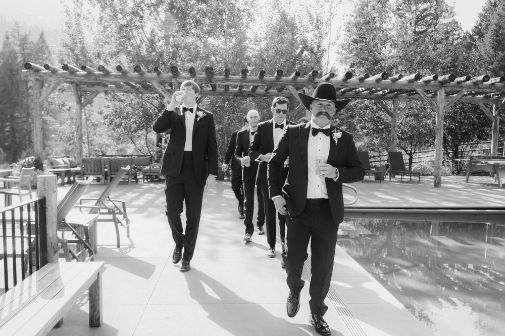 A line of formally dressed groomsmen walk in single file along a garden pathway on a sunny day.