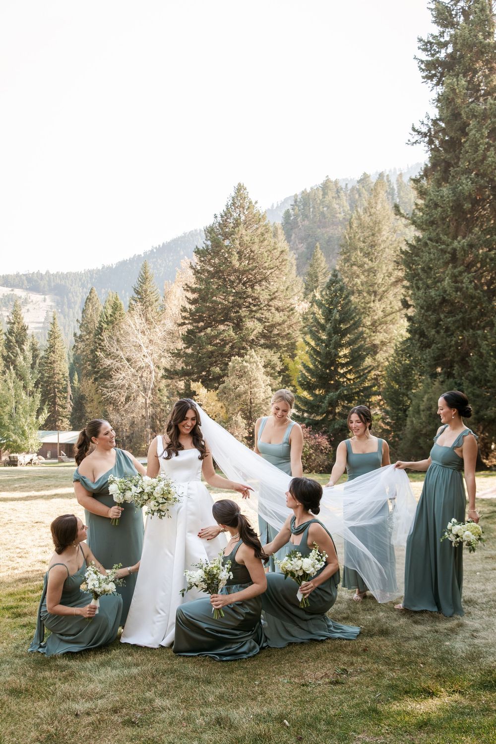 Bridesmaids in sage dresses gather around bride for a playful photo in a mountain meadow setting.