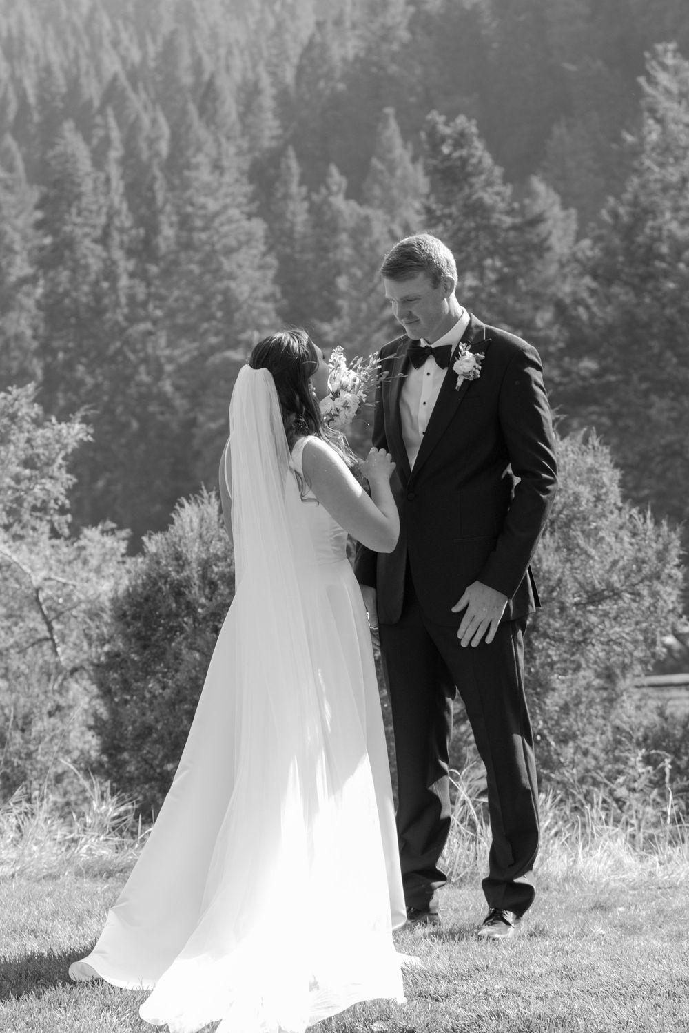 Black and white outdoor wedding photograph with couple sharing an intimate moment against a backdrop of evergreen forest.