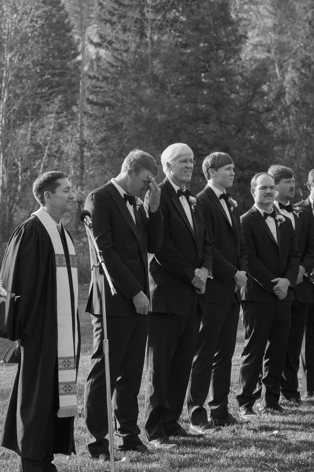 A line of groomsmen in formal black tuxedos and bow ties stand beside an officiant in robes outdoors near evergreen trees.