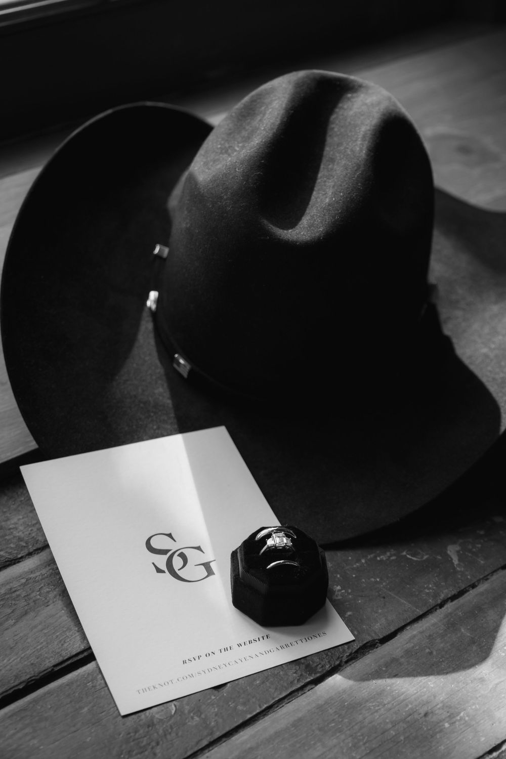 A black cowboy hat rests next to a wedding ring and invitation card in an elegant black and white composition.