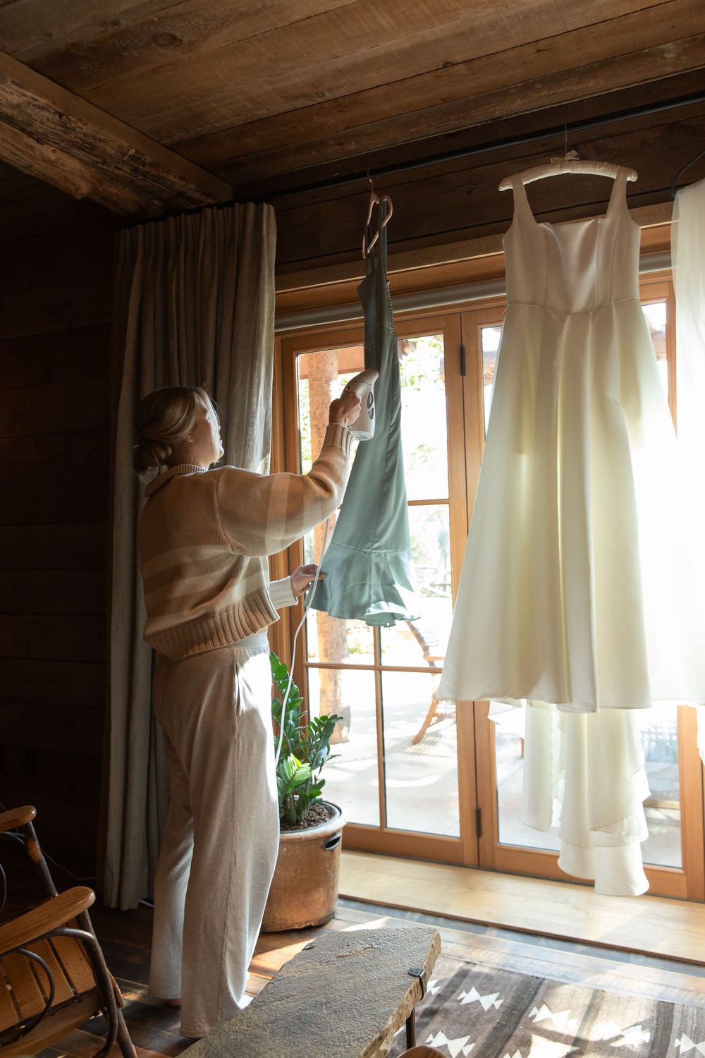 A person wearing a towel hangs white linen curtains on window rods in a rustic wooden cabin room.